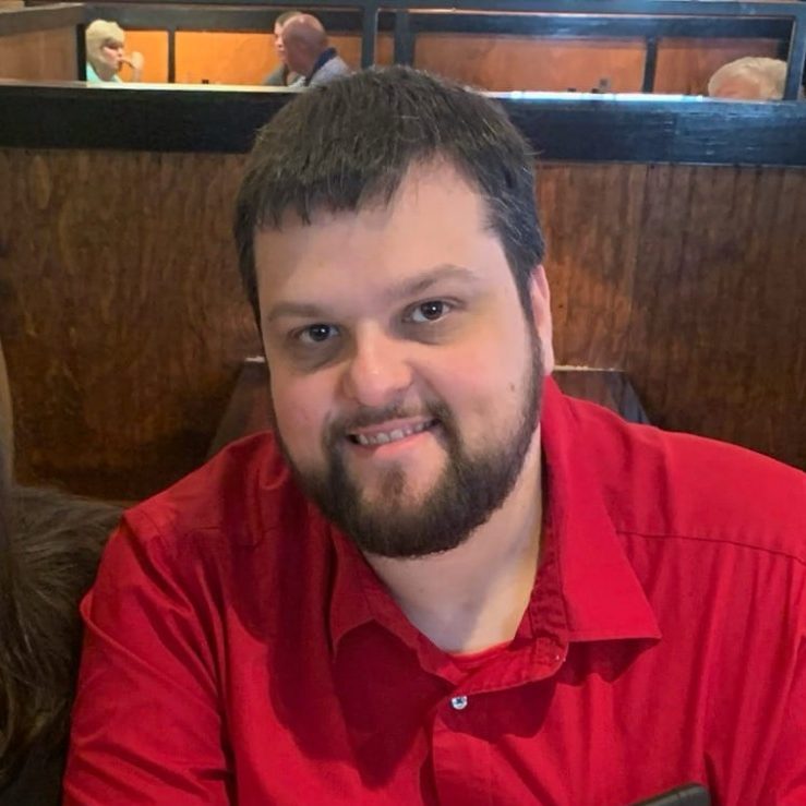 A man with a beard smiles, wearing a red shirt, seated in a restaurant.